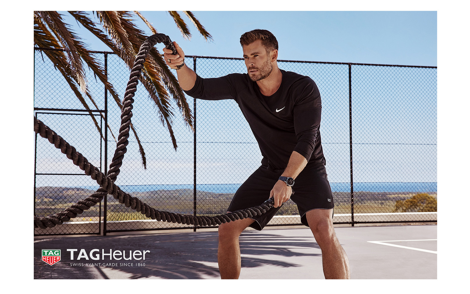 Adult male with black long sleeve shirt, shorts and watch, holding heavy ropes, and looking away at an outdoor fitness court.