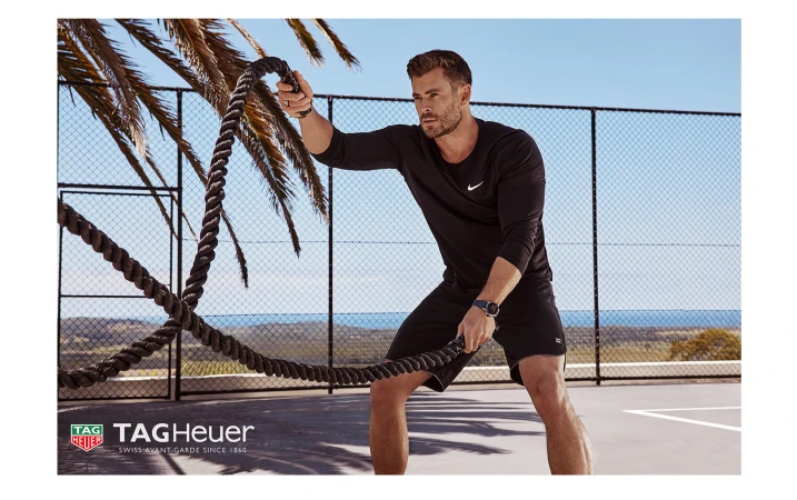 Adult male with black long sleeve shirt, shorts and watch, holding heavy ropes, and looking away at an outdoor fitness court.