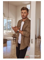 Chris Hemsworth leaning against a white doorway wearing a brown unbuttoned shirt and silver watch, in a bright domestic interior.