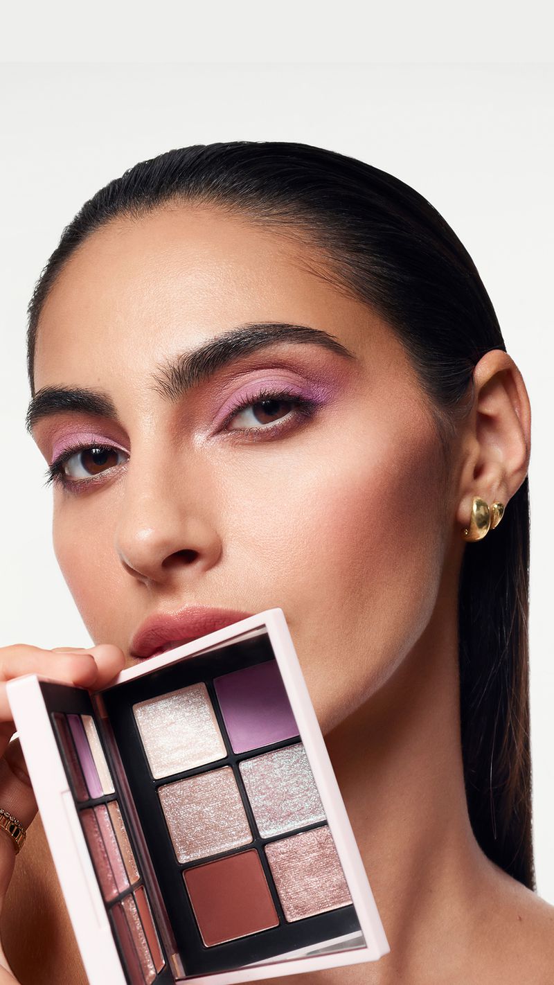 A woman with pink eye shadow holds a makeup palette in a close-up beauty shot against a white background. Prominent makeup and gold earrings.