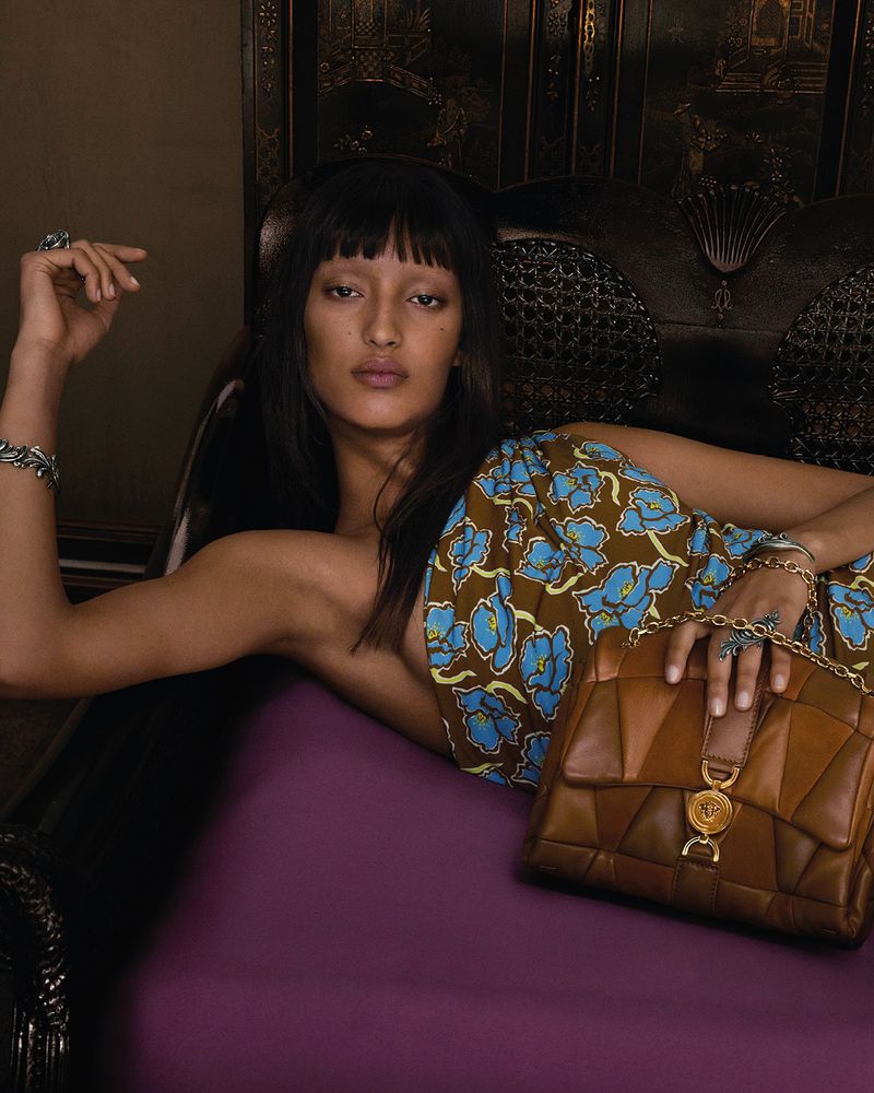 A brunette woman with bangs is lying on a purple surface with a brown leather purse in an indoor setting with dark background.