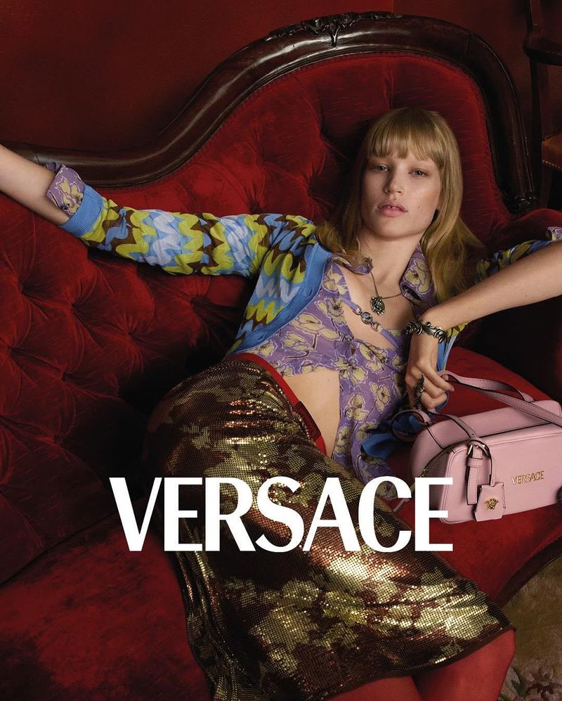 A woman lounges on a red velvet chaise wearing a Versace top and skirt, next to a pink Versace purse. The brand name is in text overlay.