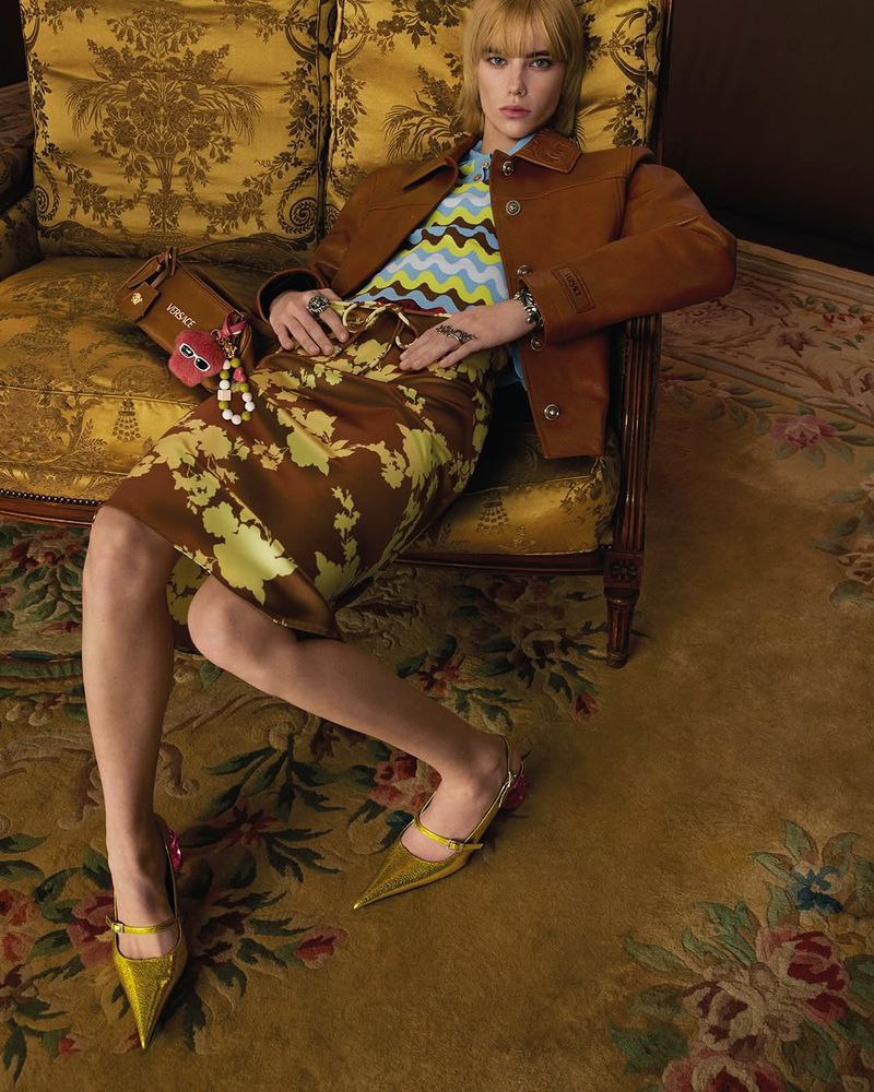Indoor fashion shot of a young woman in a floral skirt, brown jacket and gold shoes sitting on an ornate golden chair holding a purse.
