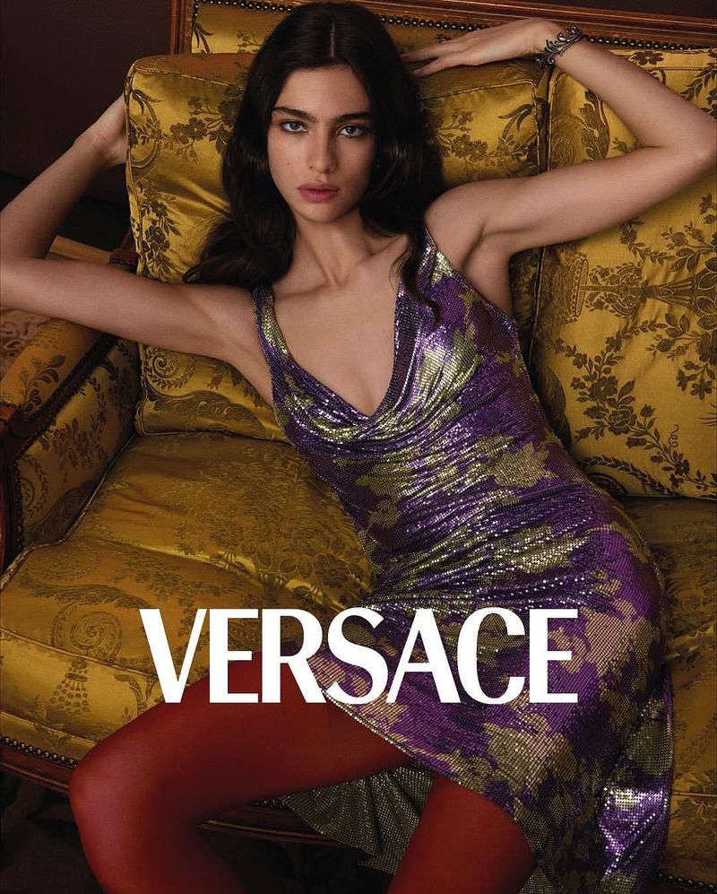 A brunette fashion model lounges on a yellow ornate couch wearing a shimmering purple dress and red tights, with the Versace logo.