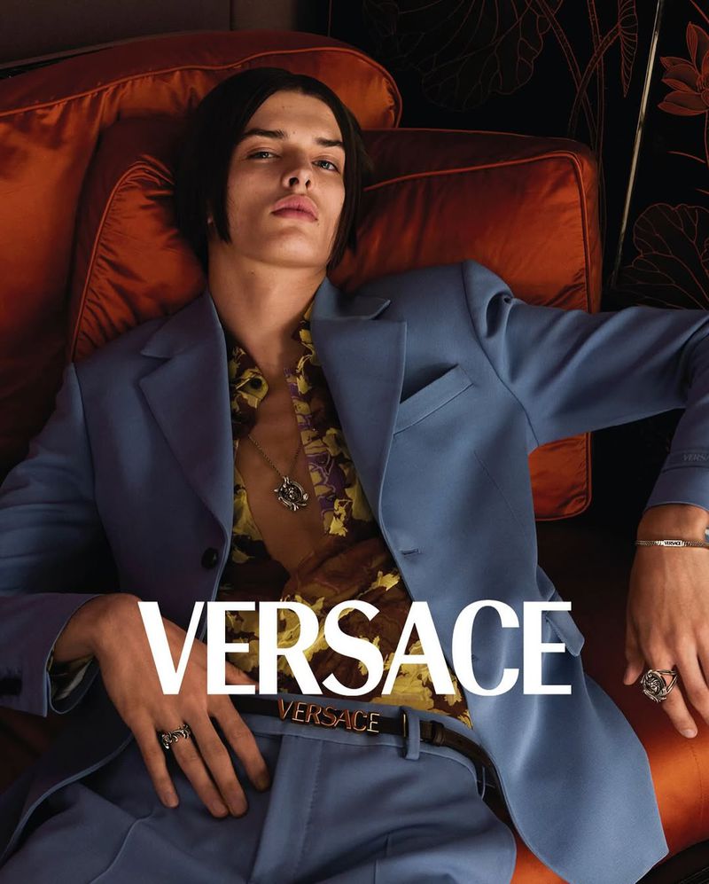 A fair-skinned young adult male model reclines on an orange cushion, showcasing a light blue suit and unbuttoned floral shirt, with the Versace logo visible.
