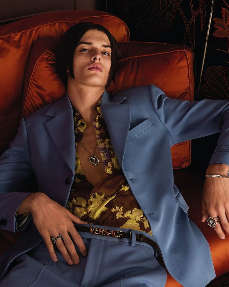 Male fashion model in a light blue suit and floral shirt reclines on an orange cushion in a close up fashion studio portrait.