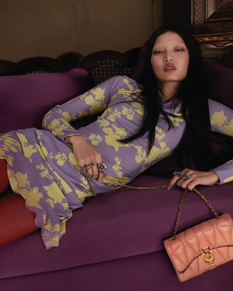 A woman with dark hair reclines on a purple sofa wearing a floral-print dress, holding a light pink purse with a gold chain strap.