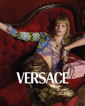 Model reclining on a plush red velvet sofa wearing a patterned top and sequined skirt with a pink handbag beside her.