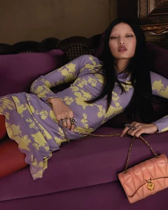 Model reclining on a purple velvet sofa wearing a lavender and yellow floral dress and holding a pink quilted chain handbag.