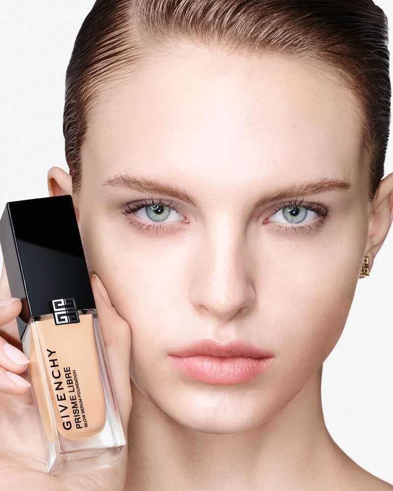 Close-up of a fair-skinned adult woman with a neutral expression holding a bottle of Givenchy Prisme Libre Glow Serum Foundation.