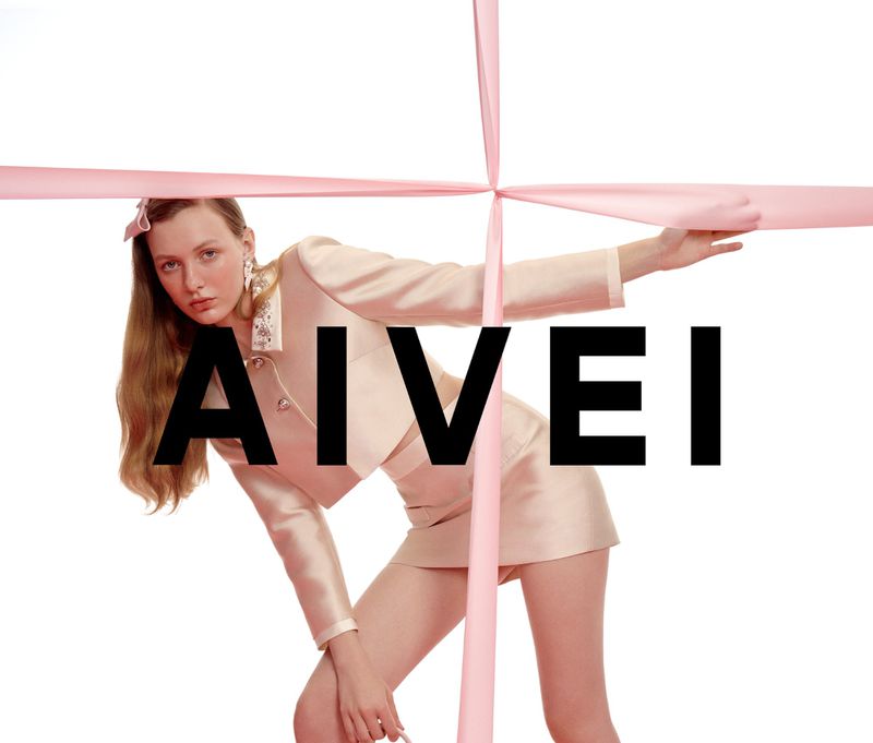 Fashion model in light pink skirt suit posing with pink ribbon. Studio shot against white background. Bold black AIVEI text overlay.
