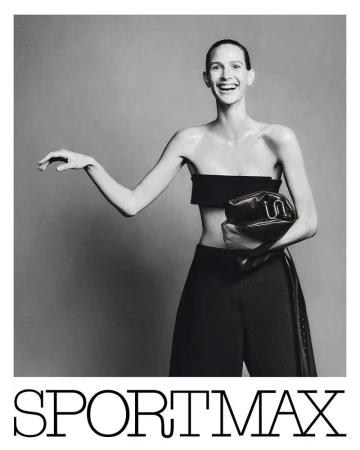 Model in black bandeau top and trousers smiling while holding a handbag against a plain grey studio backdrop.