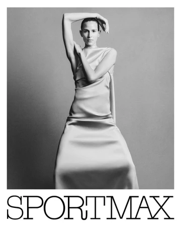 Black and white fashion shot of adult woman in a dress with arms raised against neutral backdrop with Sportmax logo at the bottom.