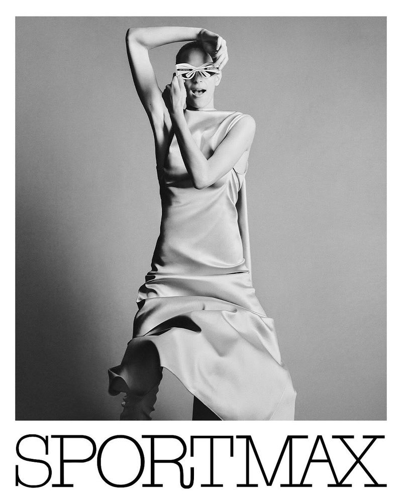 Editorial fashion shot of a bald female model in a satin dress and decorative glasses posing in a photo studio against a gray backdrop.