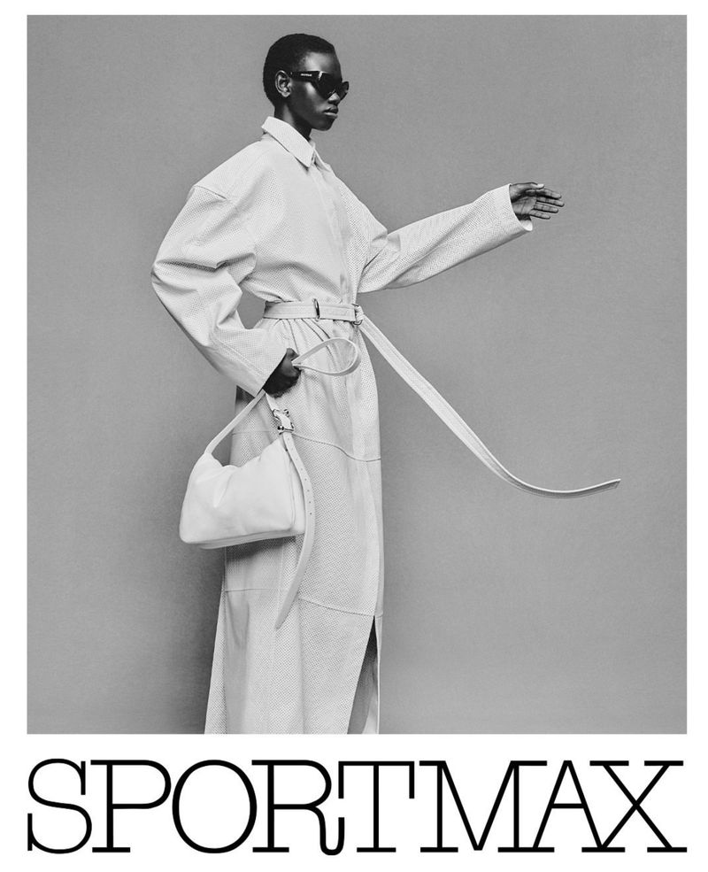 Black and white shot of a black female fashion model wearing a belted coat and black sunglasses, holding a purse in studio.