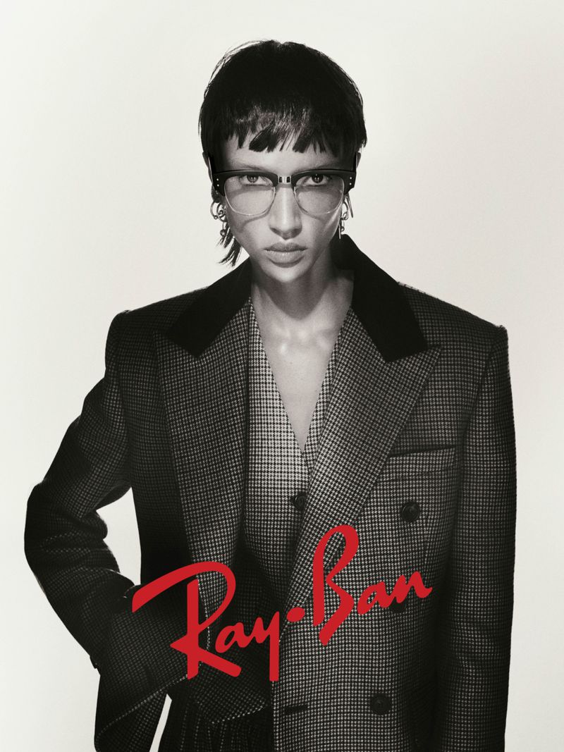 Fashion portrait of a woman wearing glasses and a double-breasted suit jacket, posing against a white background. Ray-Ban logo visible.