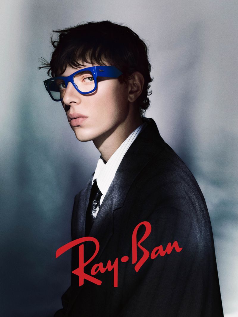 Man in a suit poses indoors, wearing blue-framed Ray-Ban eyeglasses; the red Ray-Ban logo is in the foreground.
