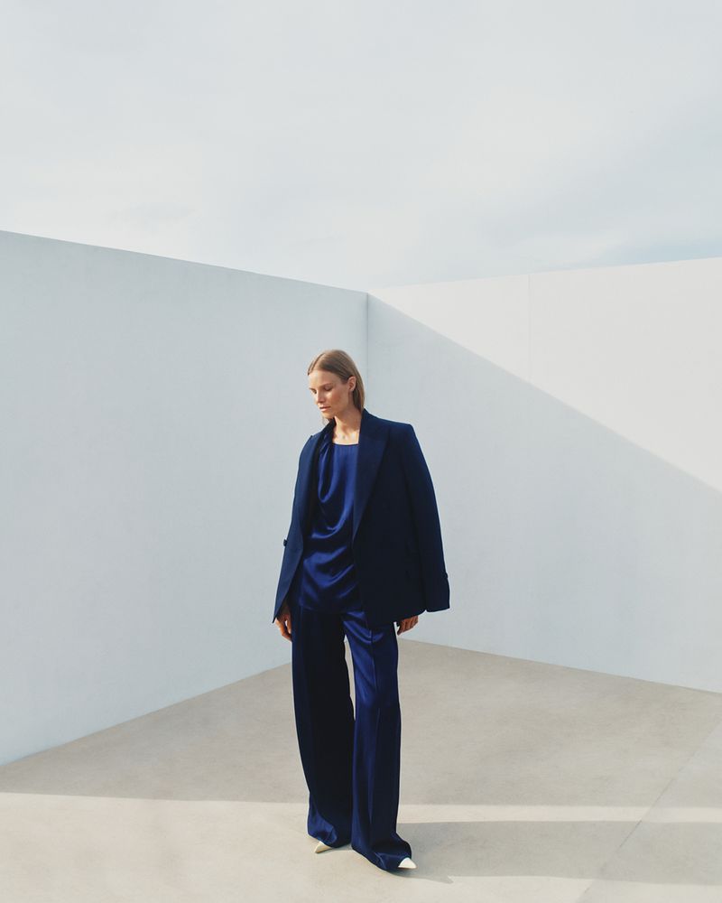 A woman models an oversized navy blue blazer and satin pantsuit against a minimalist white wall backdrop, showcasing modern fashion.