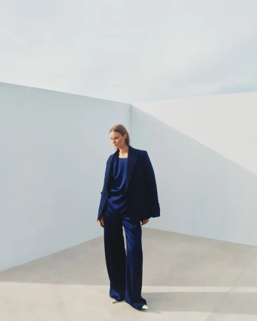 A woman models an oversized navy blue blazer and satin pantsuit against a minimalist white wall backdrop, showcasing modern fashion.