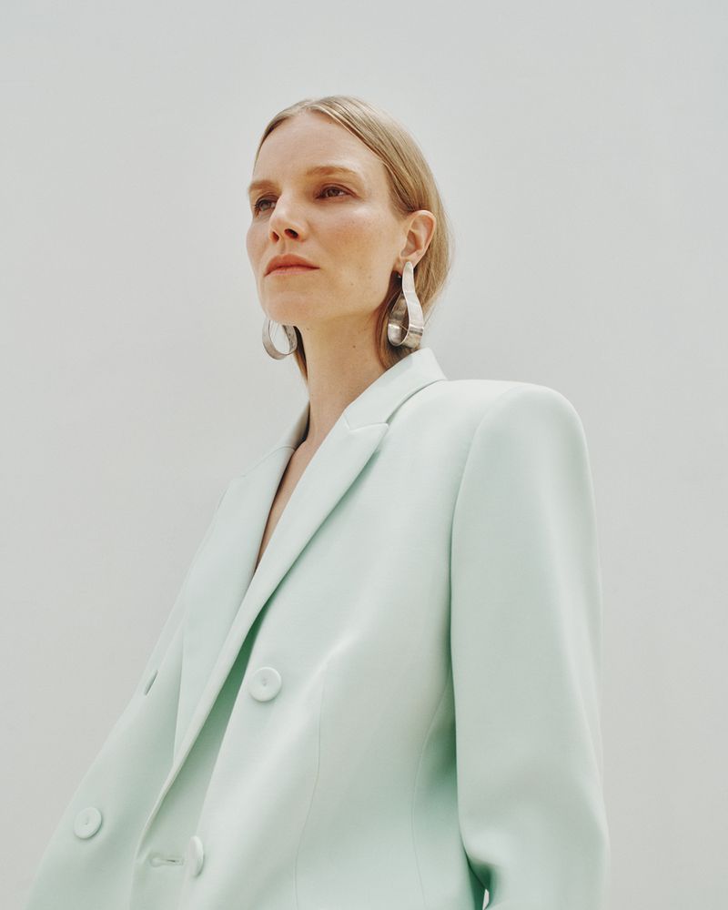 Woman in a light mint green blazer looking up with silver earrings against a white background in a fashion portrait.