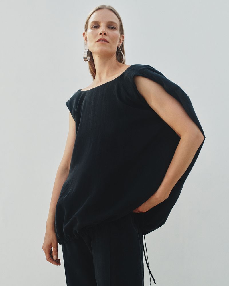 Fashion portrait of an adult woman in a black top and pants against a plain background, accessorized with hoop earrings. Neutral studio lighting.