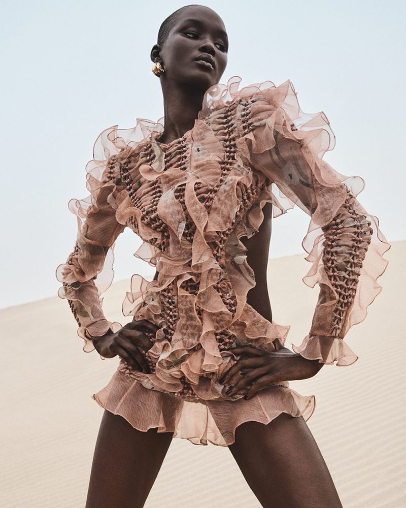 An adult female with short hair poses in a ruffled dress outdoors against a sandy landscape and bright sky in a fashion setting.