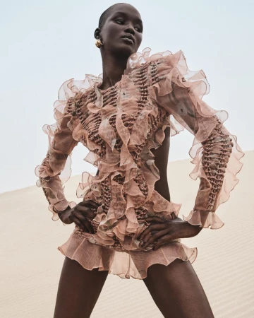 An adult female with short hair poses in a ruffled dress outdoors against a sandy landscape and bright sky in a fashion setting.
