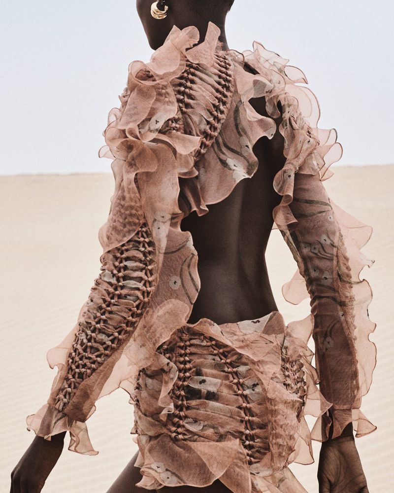 Back view of a dark-skinned woman in a sand dune landscape, wearing a peach-colored ruffled dress. The photo showcases fashion and nature.