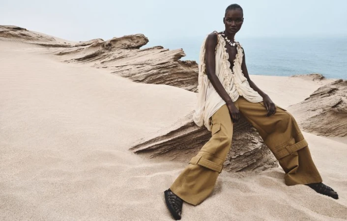 A dark-skinned adult female model poses on a sand dune wearing a flowing white top and wide tan pants, with a blue ocean backdrop.