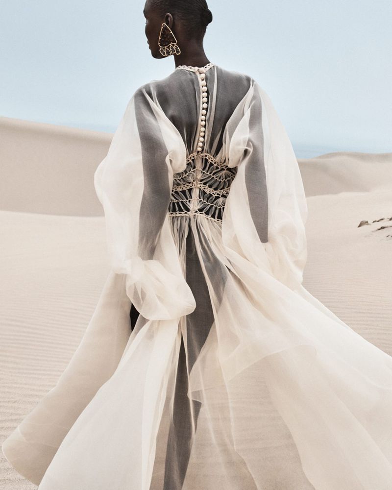 Back view of an adult fashion model in a flowing white gown with large earrings in a bright, pale desert landscape under a light blue sky.
