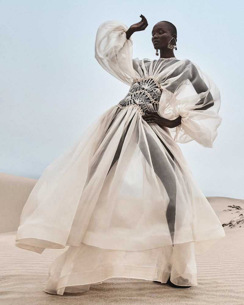 Fashion shot of Black female model in sheer, white gown with ornate corset, posing on desert sand dunes under a blue sky. High-fashion desert editorial.