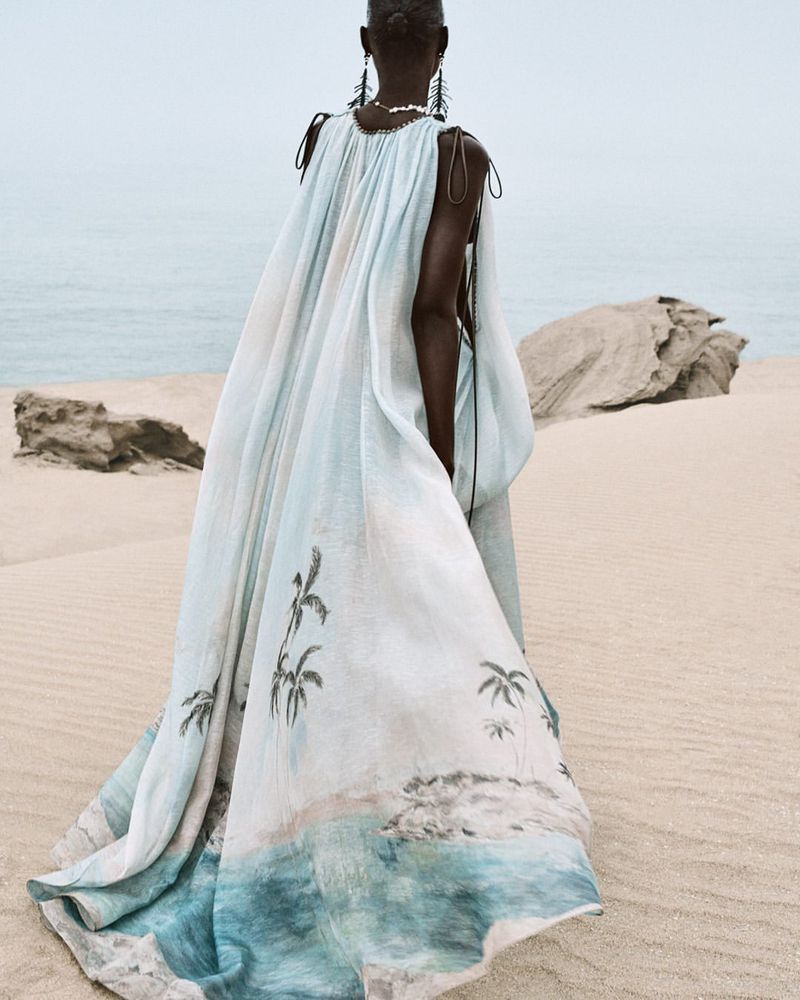 Model wears long, flowing dress with a tropical beach print on a sandy beach with an ocean view, creating a fashionable coastal scene.