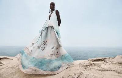 Model Abény Nhial wearing a flowing printed dress against an ocean and sand dune background for Zimmermann S/S 2025 Campaign
