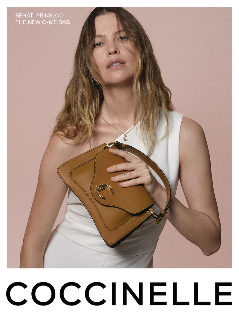 Behati Prinsloo holding a brown leather shoulder bag wearing a white dress against a solid pink backdrop for Coccinelle