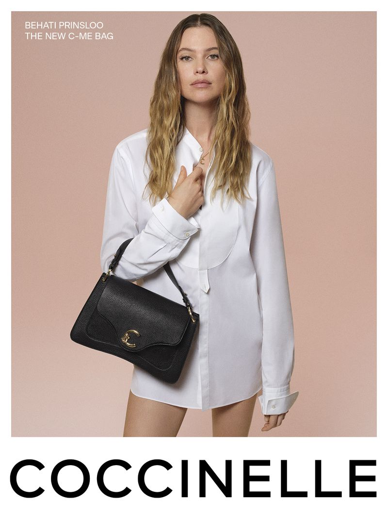 Fashion shot of a woman with long hair in a white shirt, holding a black Coccinelle handbag against a light peach background.