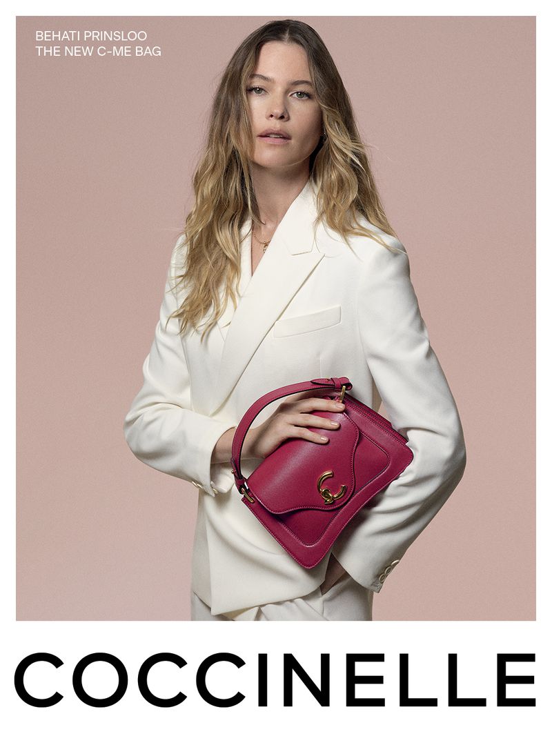 Fashion shot: adult woman in a white suit holding a red leather handbag against a pink background in a studio setting for Coccinelle.