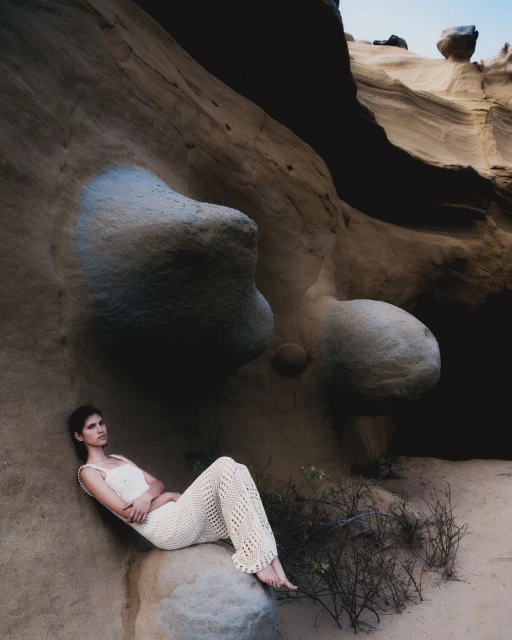 Woman in cream crochet outfit reclines on a boulder inside a canyon with tan rock formations under a hazy blue sky.