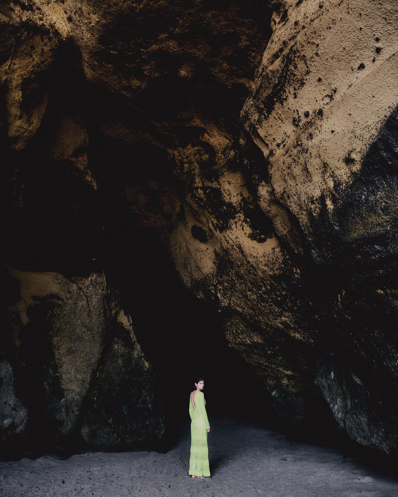 A woman in a lime green dress stands in a large dark cave, the opening is visible in the background and the cave floor is sandy.