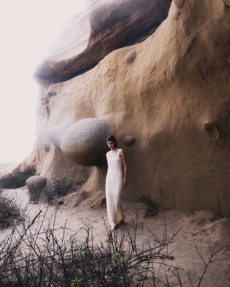 An adult female model in a long white crochet dress poses outdoors near a tall sandstone rock formation with sparse, dry vegetation.