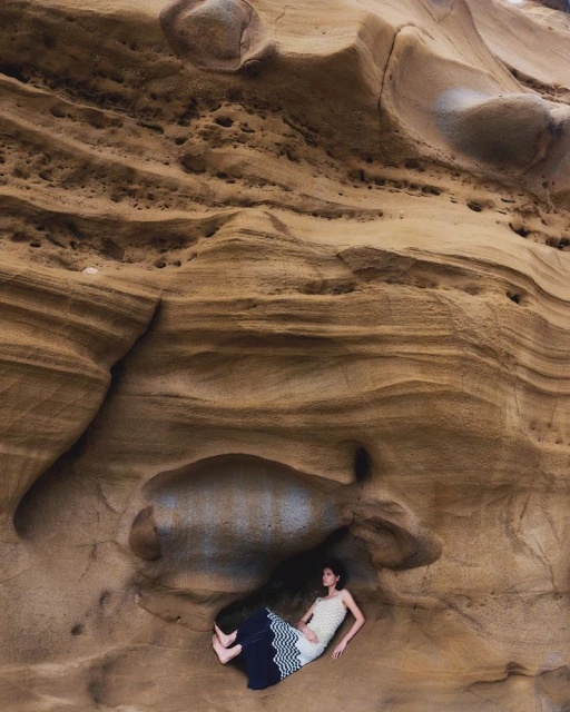 Woman in a long dress lies in a crevice in a rock formation. The neutral colors of the dress blend with the tan rock.