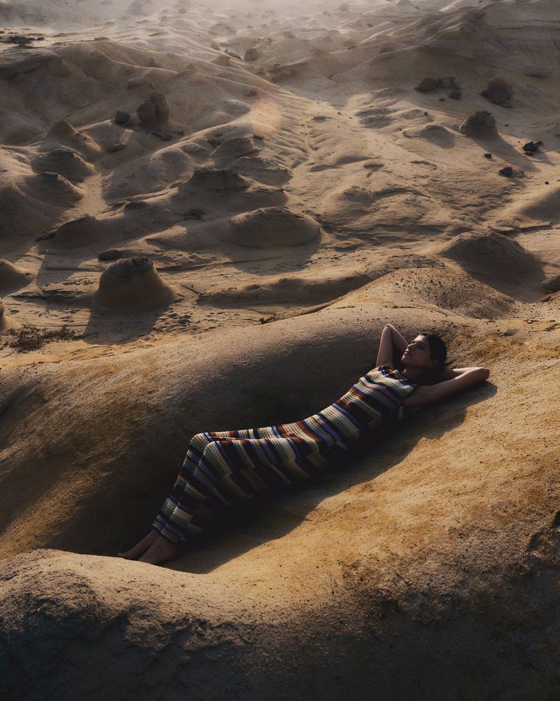 Woman in striped dress reclines on rock formations in a sun-drenched, natural environment, exuding relaxation and tranquility.