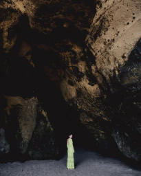 Model wearing a long lime green dress standing inside a dark rocky coastal cave opening.