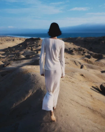 A woman wearing a cream knit top and long skirt walking barefoot across sandy coastal dunes toward the ocean.