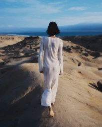 A woman wearing a cream knit top and long skirt walking barefoot across sandy coastal dunes toward the ocean.