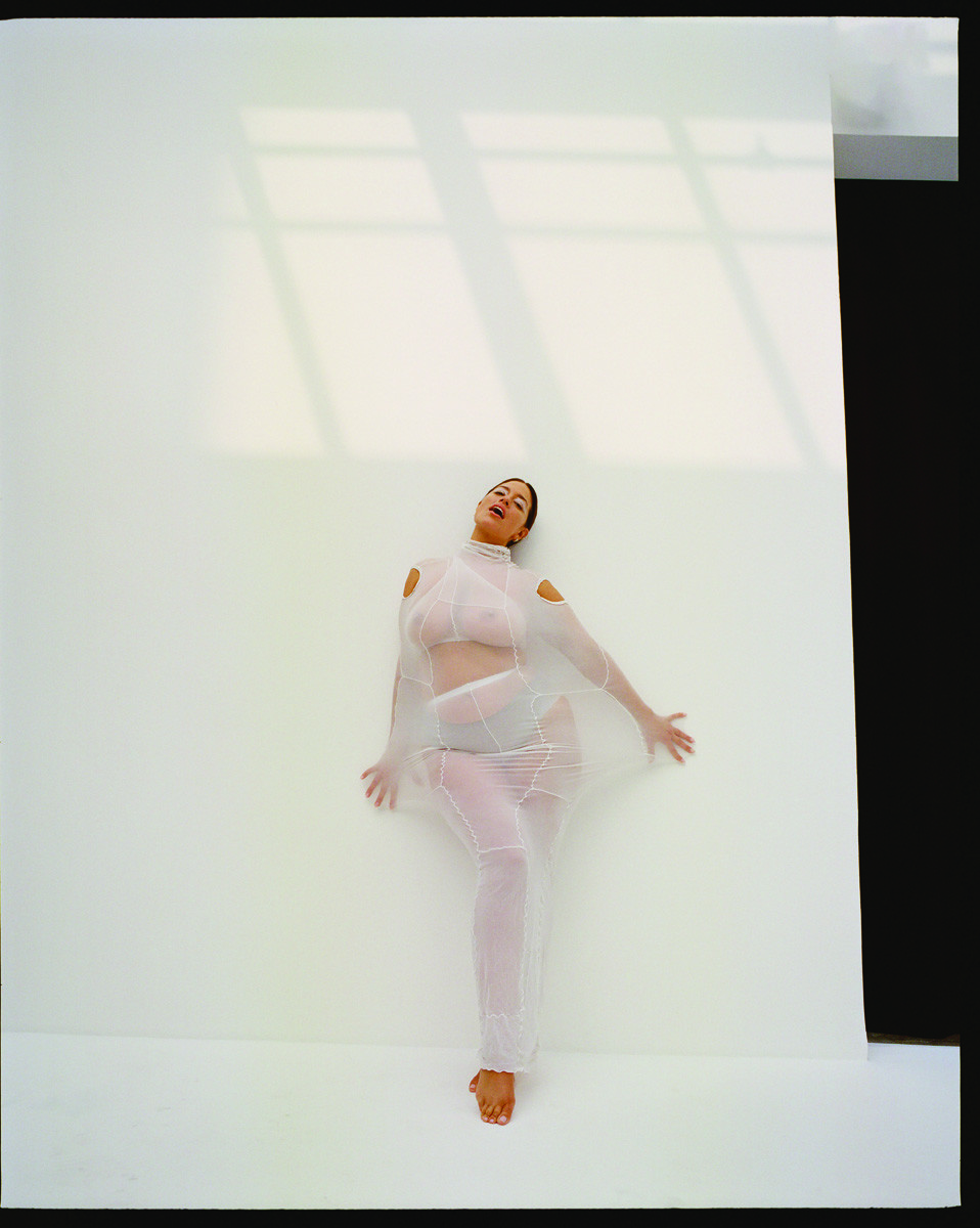 Woman poses in sheer white outfit against a white wall with window light creating shadows, evoking a modern fashion editorial.