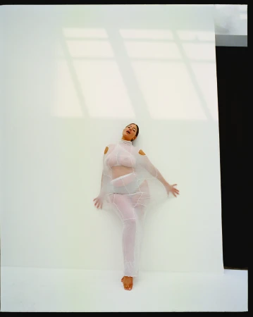 Woman poses in sheer white outfit against a white wall with window light creating shadows, evoking a modern fashion editorial.