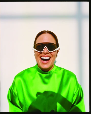 Portrait of an adult woman smiling, wearing white-rimmed sunglasses and a bright green top, indoor studio shot with shadow.