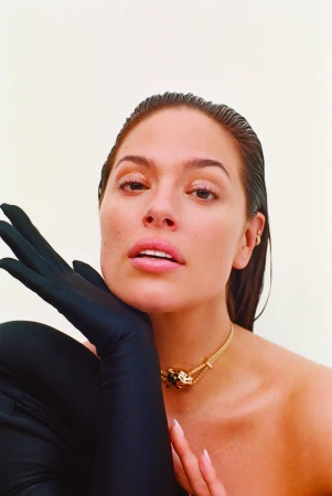 Close-up portrait of model Ashley Graham wearing black opera gloves and a gold choker necklace against a seamless light background.
