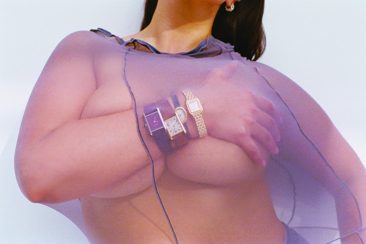 Adult female wearing sheer purple top and three wrist watches, posing against a white background in an indoor environment.