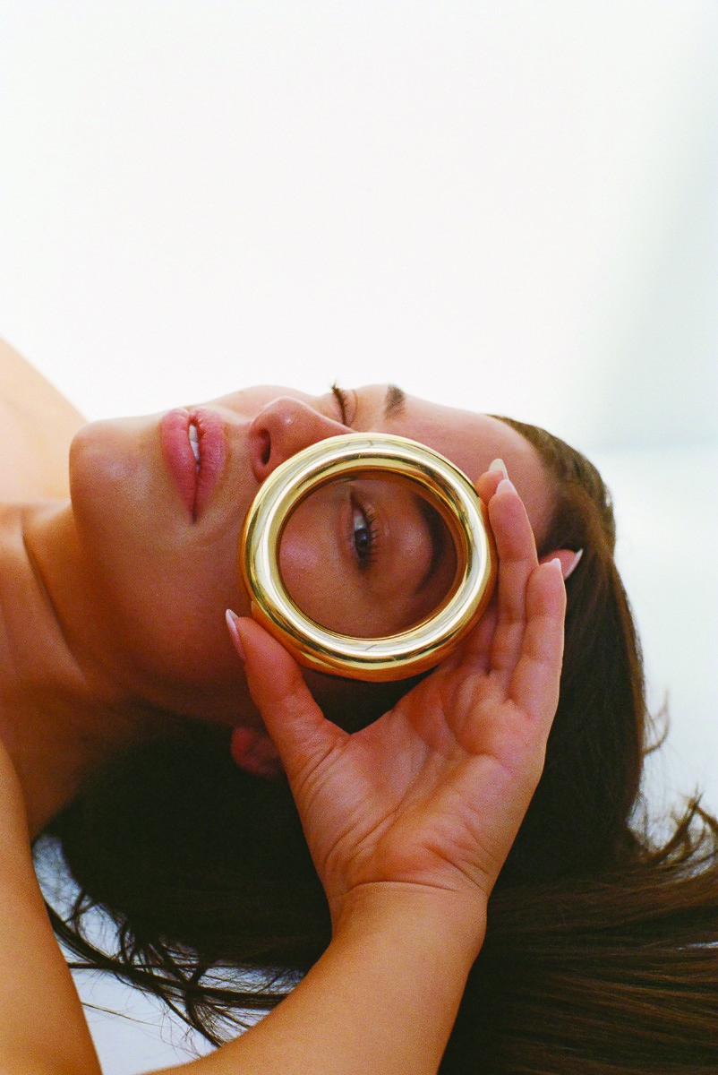 Eye-level studio shot of a woman lying down, her dark hair spread on a white surface, holding a gold bangle to her eye.